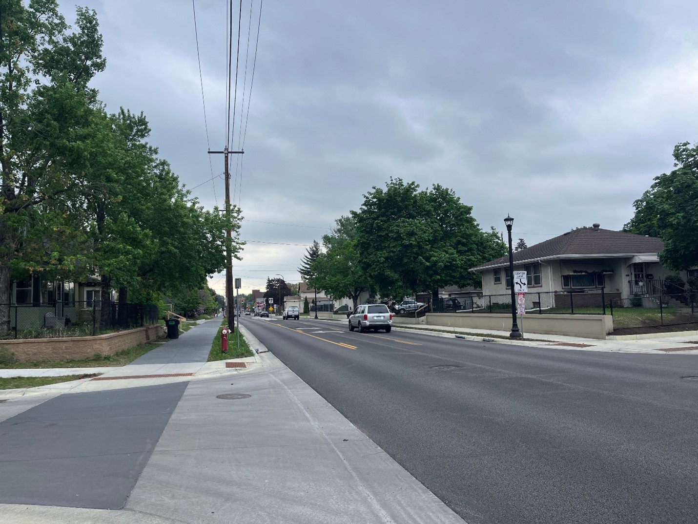 A neighborhood street showing cars on the road and a trail on the left side.