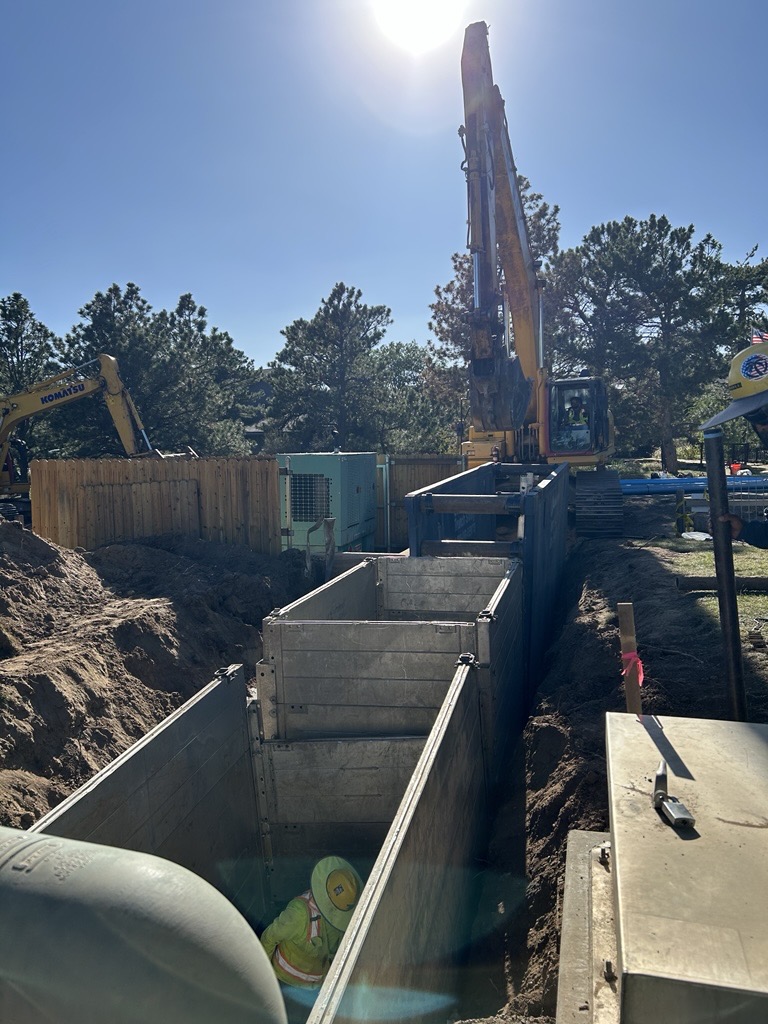Two large trench boxes support an excavated area with a work in one box and construction equipment in the background. 