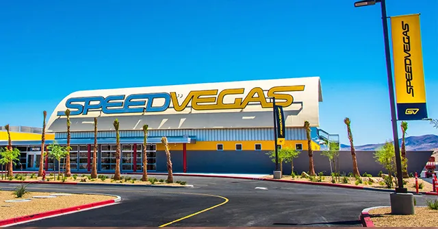 Bright, sunny day at SpeedVegas, featuring a large building with bold yellow lettering. Palm trees line the entrance, with desert mountains in the background.