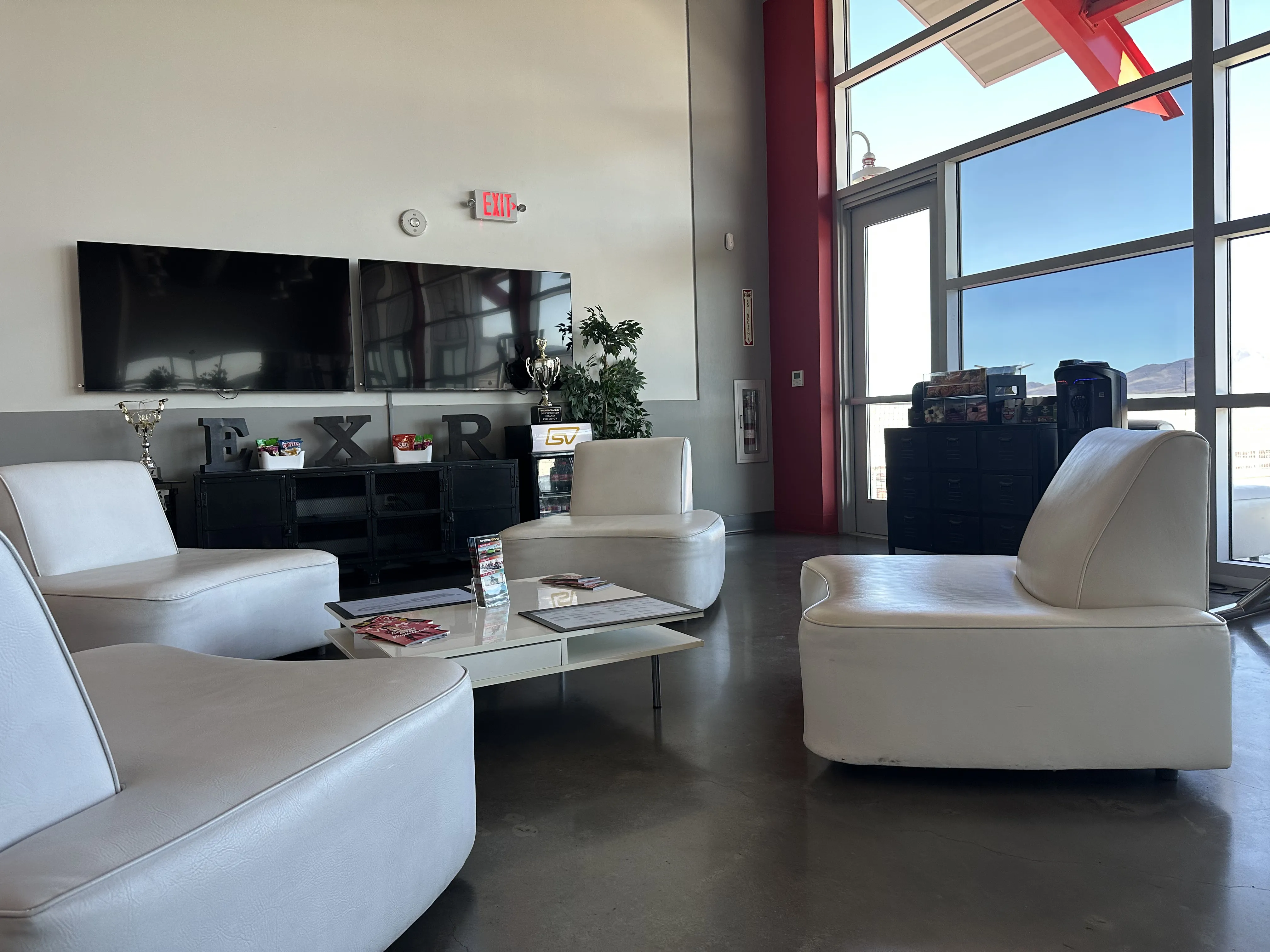 Modern lounge area with white leather chairs arranged around a glass coffee table. Two wall-mounted TVs and trophies are visible. Sunlight streams through large windows.
