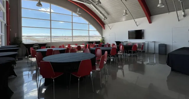 Spacious room with red chairs surrounding black round tables, large arched windows showing a clear sky. Modern, minimalistic, calm setting.