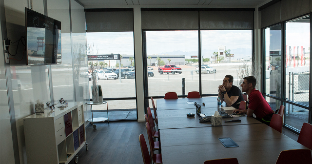 Two people sit at a conference table in a modern, glass-walled room, attentively watching a wall-mounted TV. Sunlight streams through large windows.