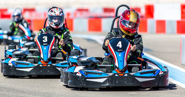 Two go-kart racers compete on a sunny track, wearing colorful helmets and suits. The scene is dynamic and competitive, with a focus on speed.