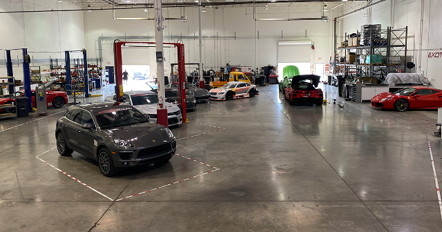 A spacious auto workshop with multiple cars, including a gray SUV in the foreground and sports cars in the middle. The area is organized and well-lit.