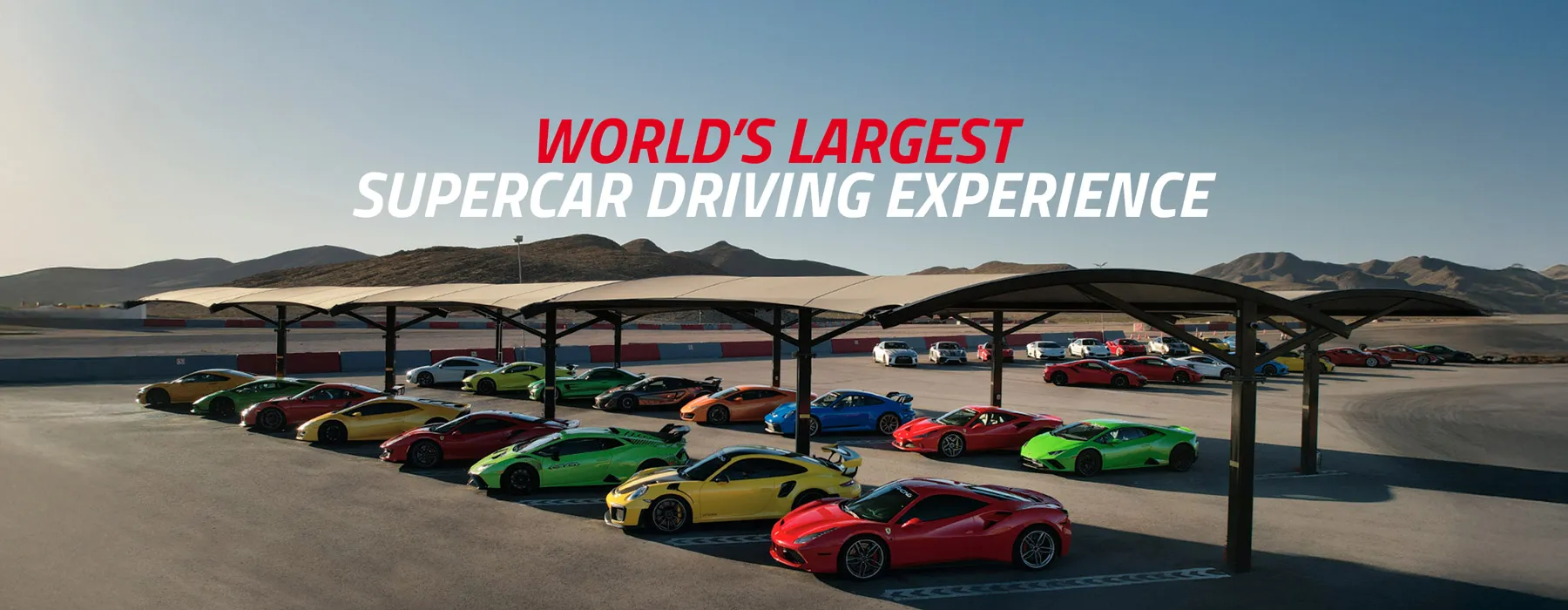 A fleet of colorful supercars under shade structures on a racetrack with mountains in the background.