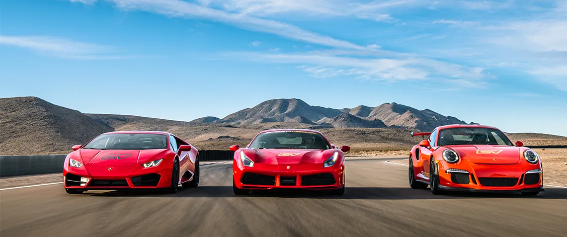 Three vibrant sports cars, two red and one orange, speed side by side on a racetrack with desert mountains in the background under a clear blue sky.