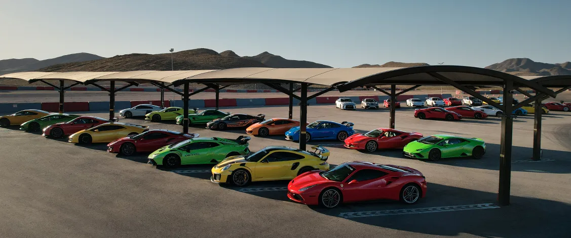 A vibrant collection of colorful sports cars, including red, green, yellow, and blue, parked under a canopy on a racetrack. Mountains visible in the background.