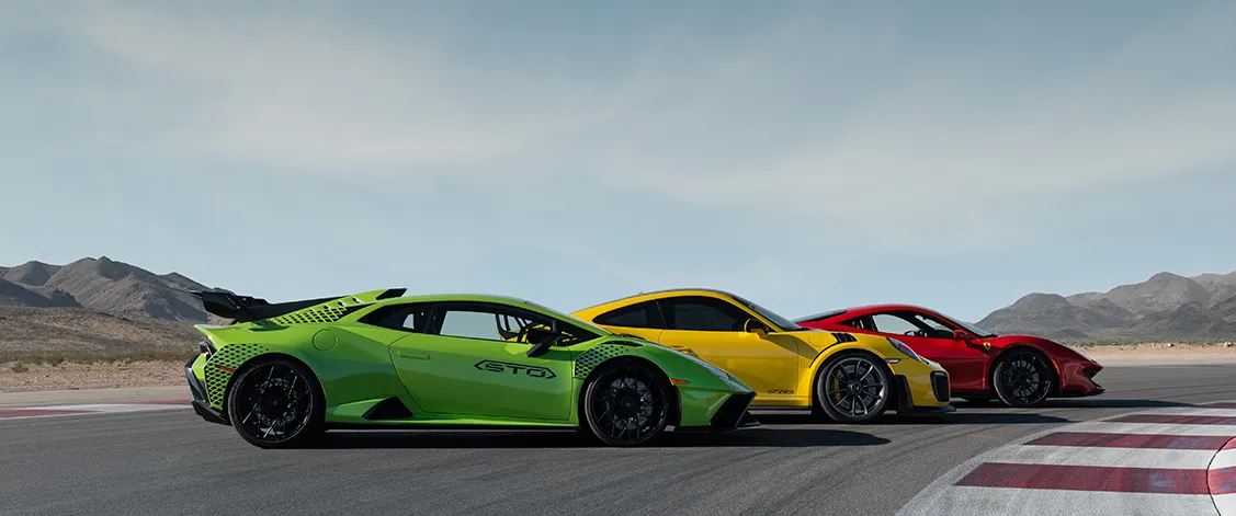 Three sleek sports cars, green, yellow, and red, line up on a racetrack under a vast blue sky, exuding speed and excitement against a desert landscape.