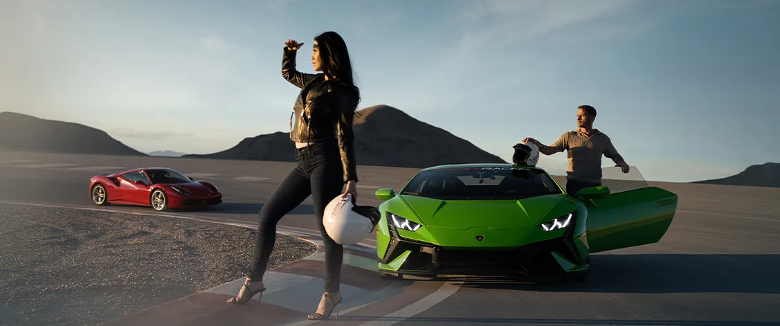 A woman in a leather jacket and high heels holds a helmet, near a green sports car on a sunlit racetrack. A red sports car is visible in the background.