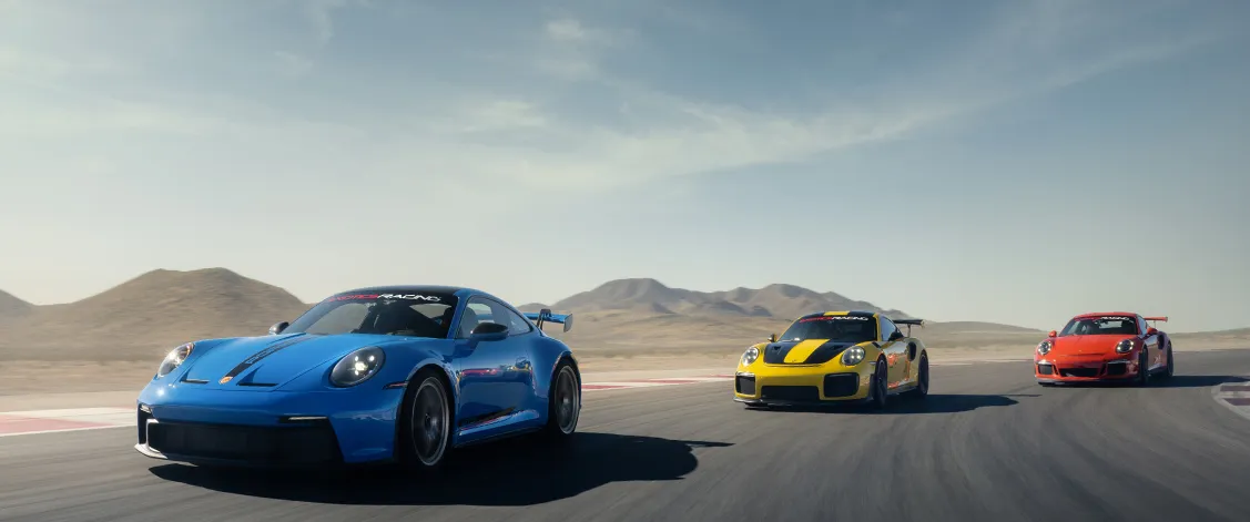 Three sports cars, one blue, one yellow, and one red, race on a desert track under a clear sky, creating a dynamic and exhilarating scene.