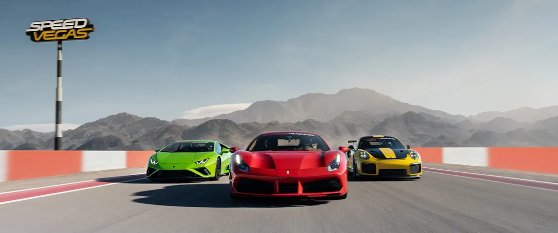 Three sports cars race side by side on a track against a mountainous backdrop. From left to right: green, red, yellow. 'Speed Vegas' sign above.