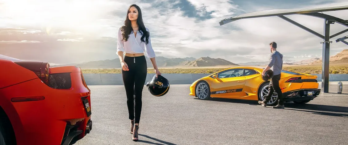 A woman holding a helmet walks confidently on a racetrack, flanked by a red car and a yellow sports car. A man stands nearby, with arid mountains in the background. The scene conveys excitement and luxury.