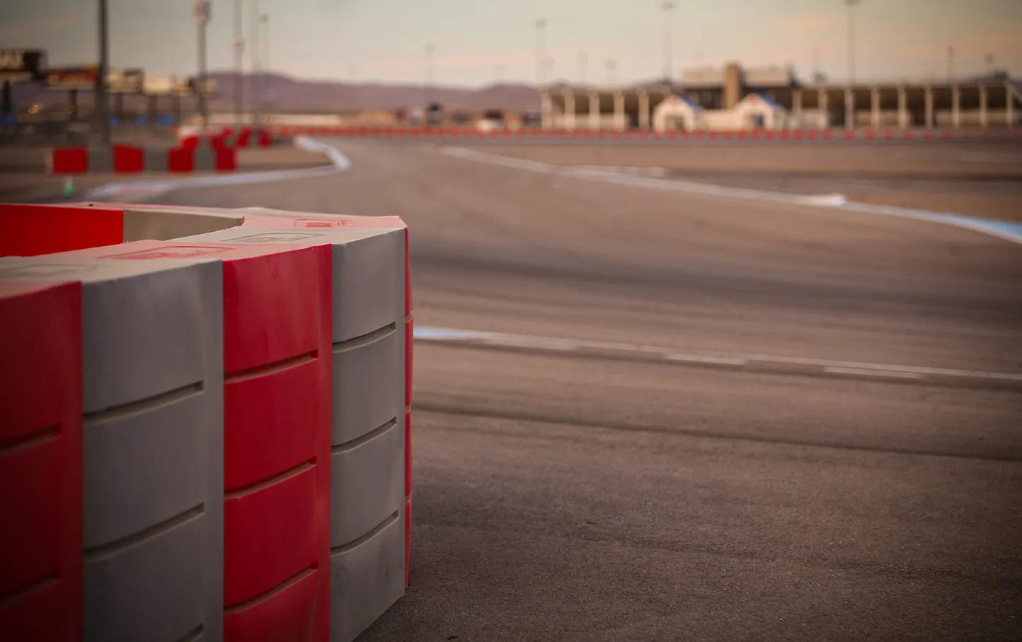 Race track curve with red and gray barriers in the foreground. Empty asphalt extends into distance under a clear sky, creating a calm atmosphere.