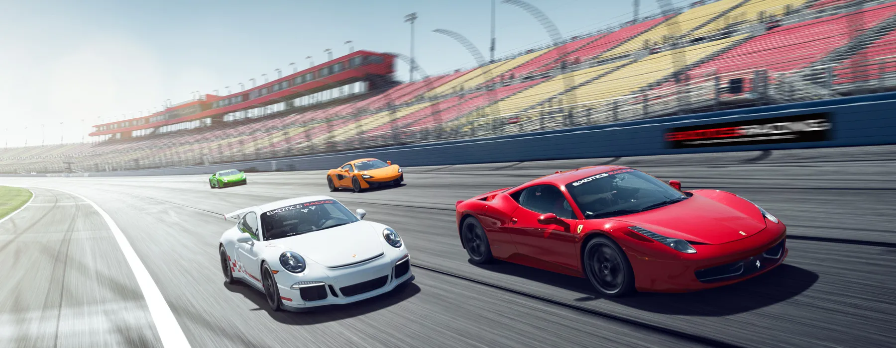 Four sports cars race on a curving track. A red car leads, followed by a white, orange, and green car. Grandstand in the background, sunny day.