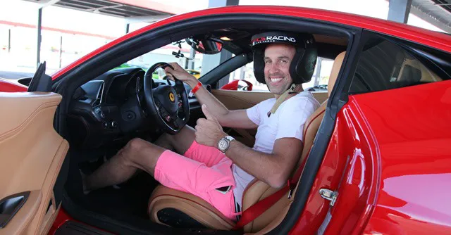 Man in a white shirt and pink shorts sits in a red sports car, smiling and giving a thumbs-up. He wears a racing helmet, conveying excitement and confidence.
