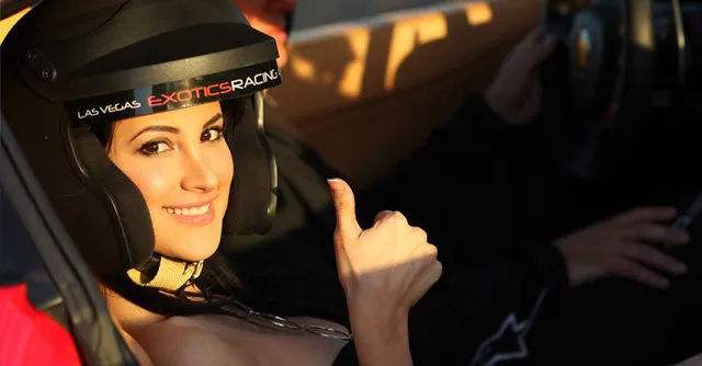 Woman in a black racing helmet sits in a car, smiling and giving a thumbs-up. The setting suggests excitement and enjoyment in a sports car environment.