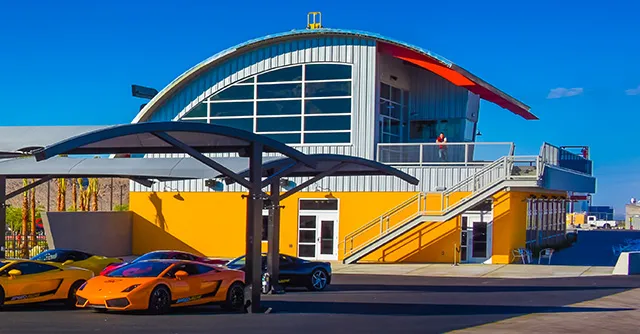 A vibrant scene featuring a modern yellow building with a curved roof under a clear blue sky. Colorful sports cars are parked in front, conveying luxury and excitement.