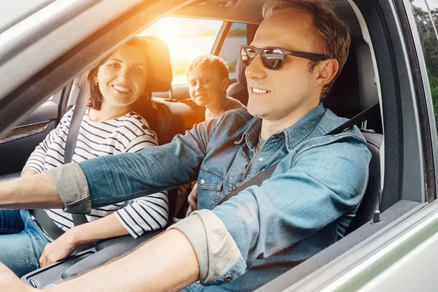A smiling family in a car, with a man driving, a woman in a striped shirt as passenger, and a child in the backseat. Sunlight streams through the windows.
