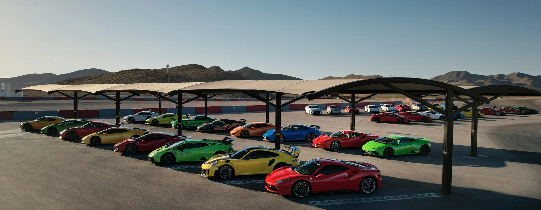 A vibrant lineup of luxury sports cars in various colors, including red, yellow, and green, parked under shade structures at a racetrack; mountains in the background.