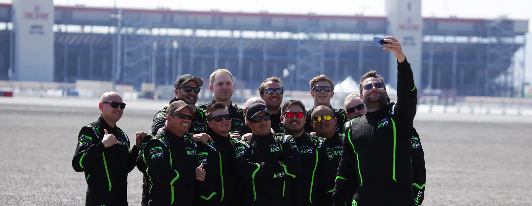 A group of 11 men in black and green racing suits pose for a selfie on a racetrack. The mood is cheerful, with a large stadium in the background.