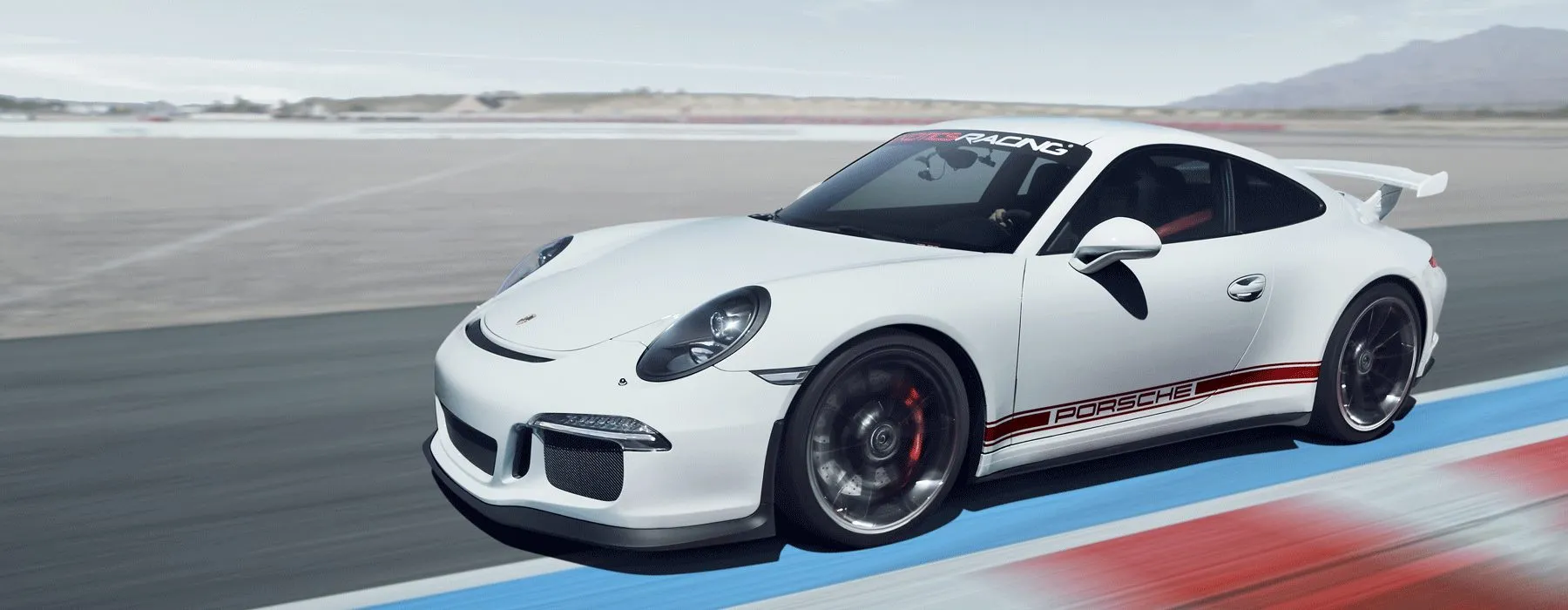 Sleek white sports car speeding on a racetrack, featuring red Porsche branding. Motion blur on wheels, mountains in the background under a clear sky.