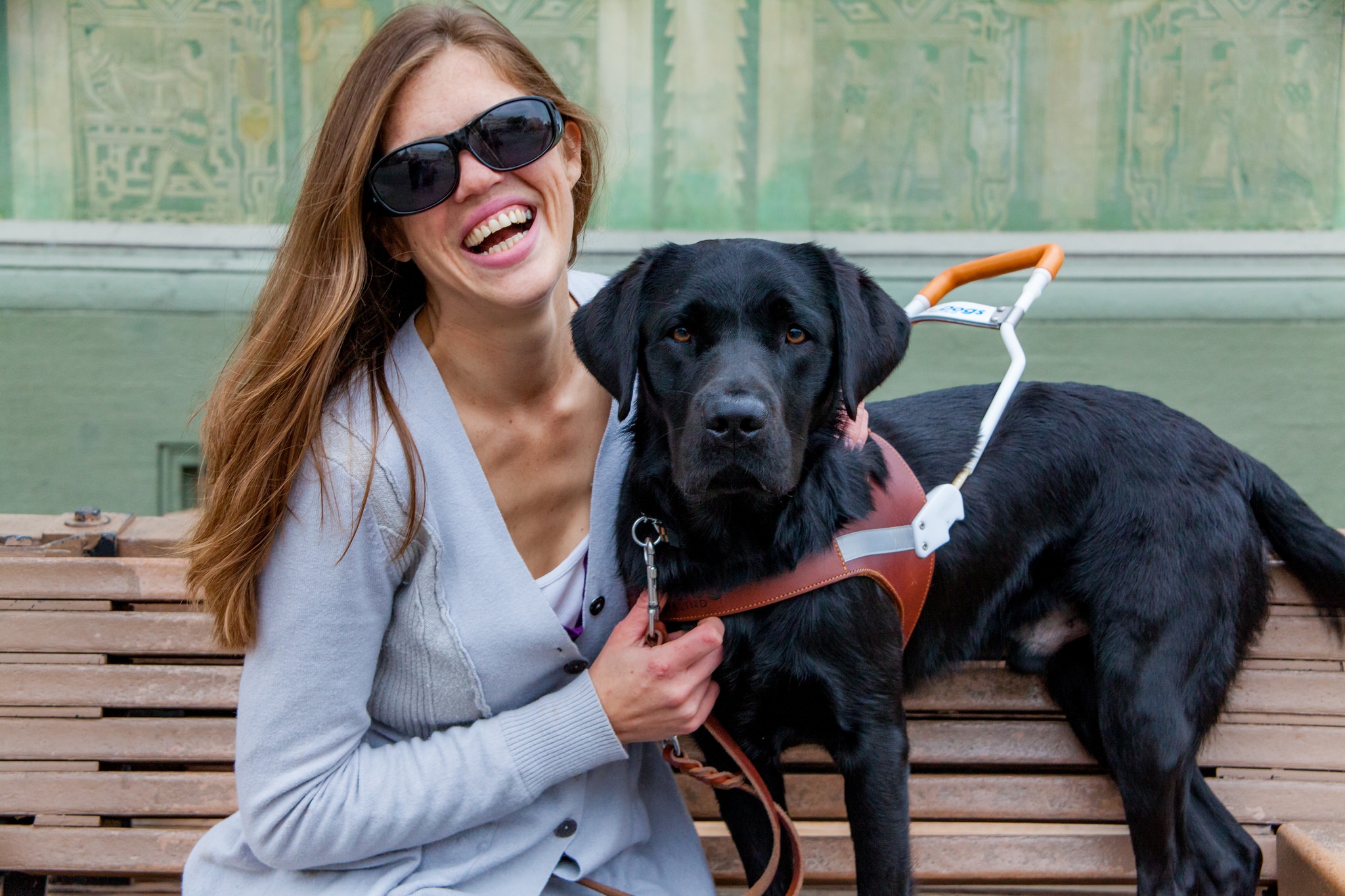 GDB alumna Dorlyn Catron smiles while sitting on a bench with her black Lab guide dog Tank. Dorlyn is wearing dark black sunglasses and a gray sweater. Tank is wearing a guide dog harness.
