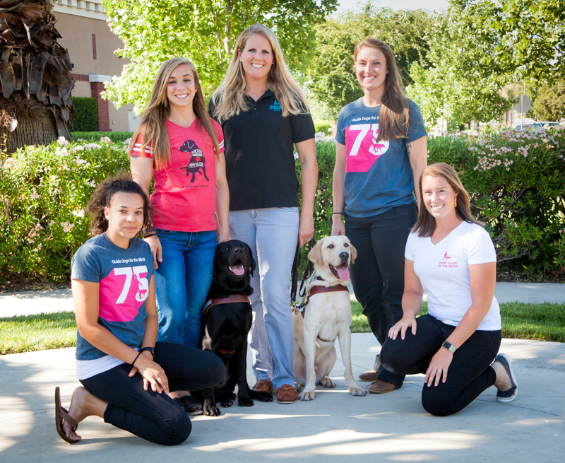 GDB's five new guide dog mobility instructors: Jessie St. Clair, Amanda Baumgartner, Palmer Dooley, Addie Yake, and Jane Allman, with two guide dogs in harness (one black Lab and one yellow Lab).
