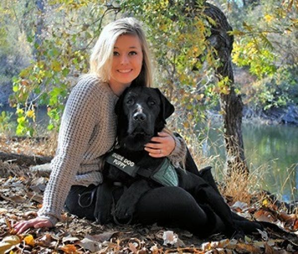 Emily sits with her black Lab guide dog puppy on the ground surrounded by leaves