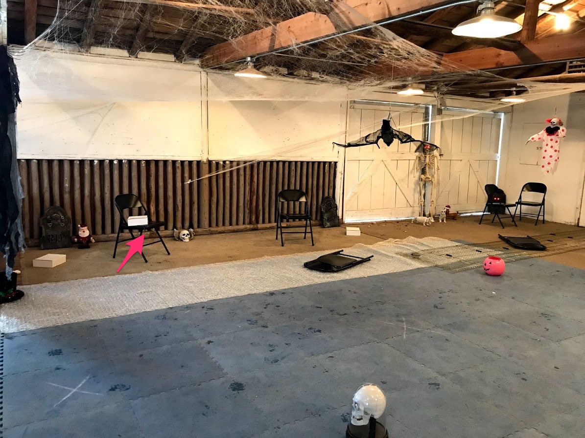 Part of a room cordoned off by bubble wrap laid on the floor. Chairs and misc halloween decorations are on one side of the bubble wrap, where the dogs would be searching.