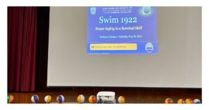 The opening slide for Swim 1922: Water Safety is a Survival Skill, presented by the Eta Nu Sigma Brooklyn Alumnae Chapter of Sigma Gamma Rho Sorority, Inc. and the USA Swimming Foundation, projected on screen at Bushwick Campus on Saturday, May 30, 2026. Colorful beach balls line the stage in front of the screen.