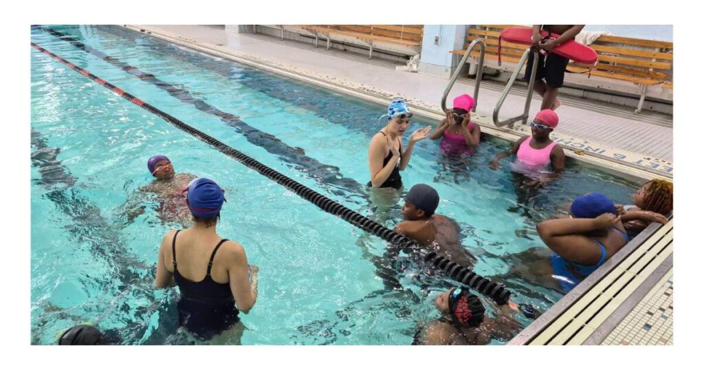 A Swim Strong Foundation instructor leads adult participants through a free swim clinic in the pool at Bushwick Campus during WaterPalooza 26, with a lifeguard on deck and multiple participants working on water skills across two lanes.