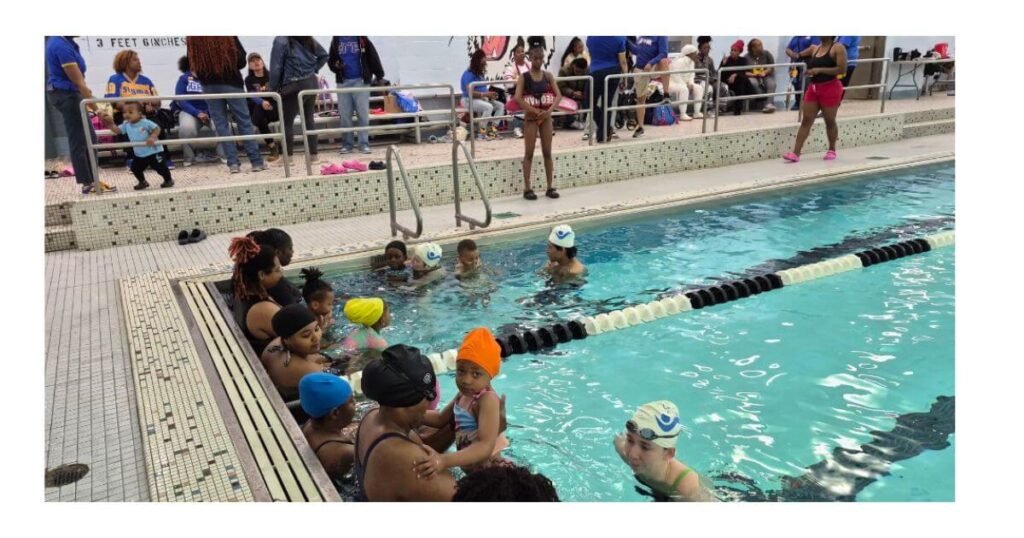 Parents and young children participate in introductory water safety and swim activities at the pool during WaterPalooza 26 at Bushwick Campus, with members of the Eta Nu Sigma Brooklyn Alumnae Chapter of Sigma Gamma Rho Sorority, Inc. cheering from the deck.