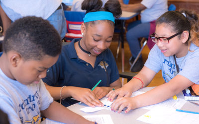 Three students collaborating on project at table, looking down at resources