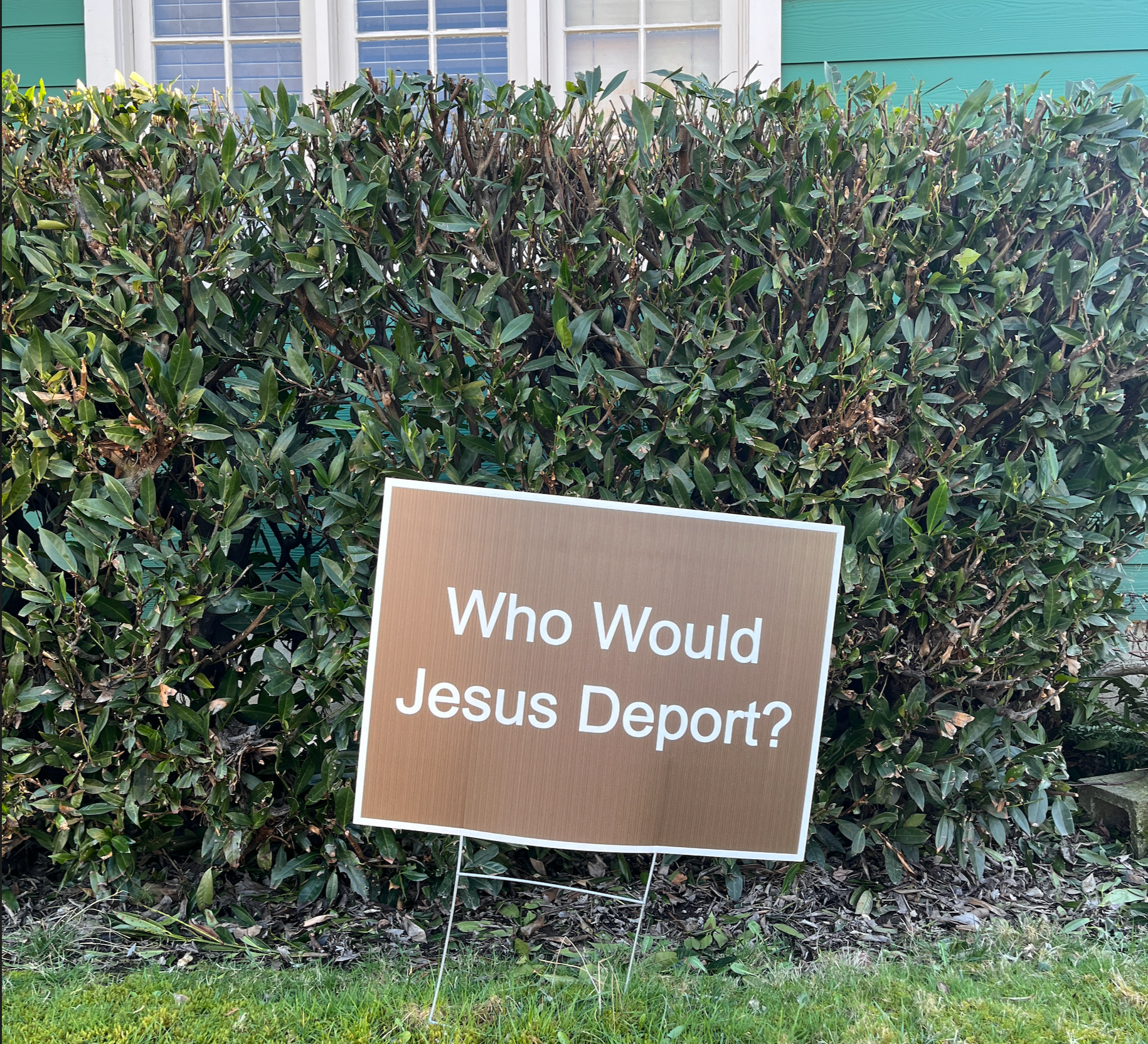 Brown yard sign with white letters in front of a green house saying, "Who Would Jesus Deport?"