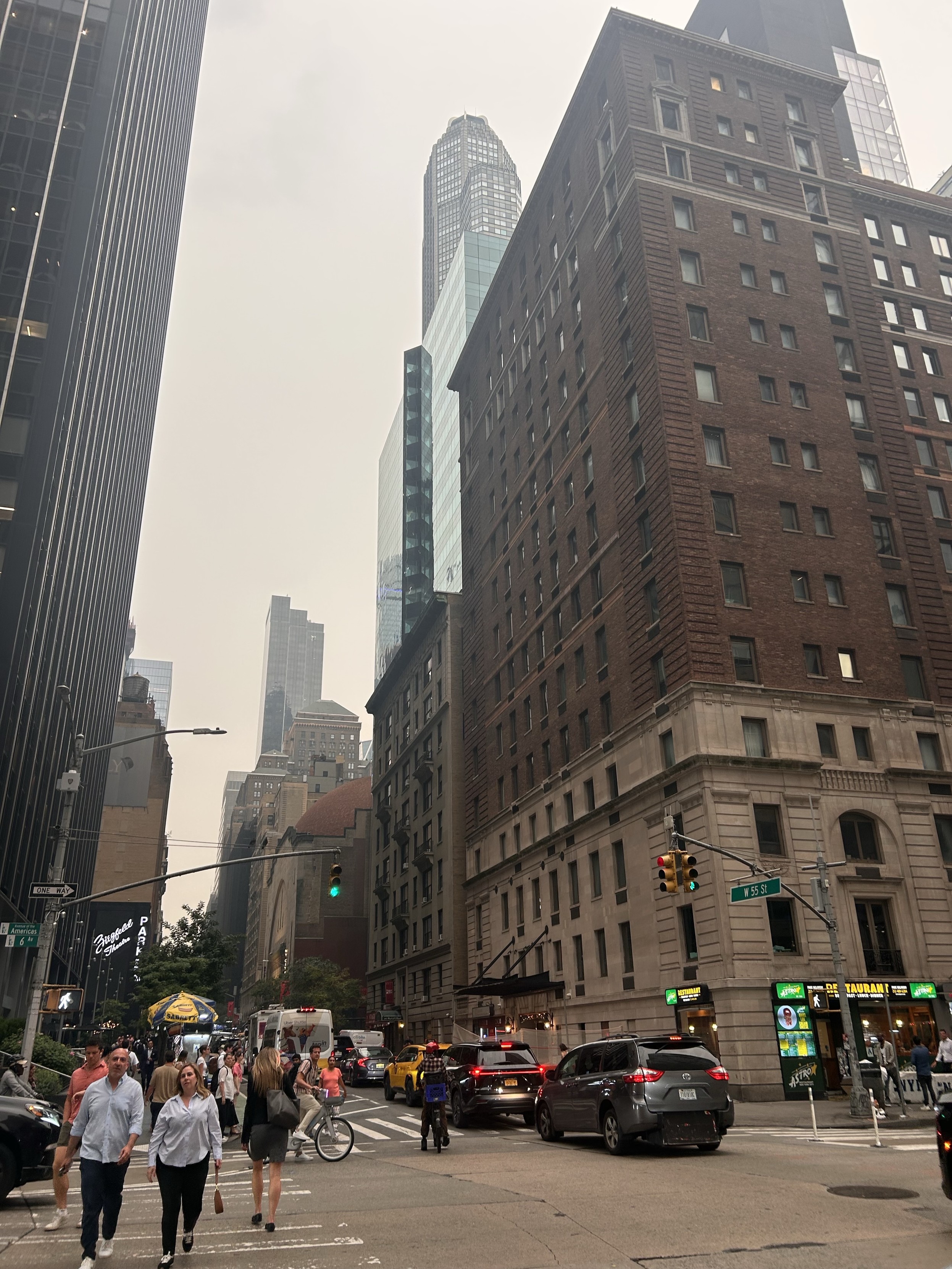 A busy urban street scene featuring pedestrians crossing the road and vehicles waiting at a traffic light. Tall buildings, including modern skyscrapers and older brick structures, are visible in the background under a hazy sky.