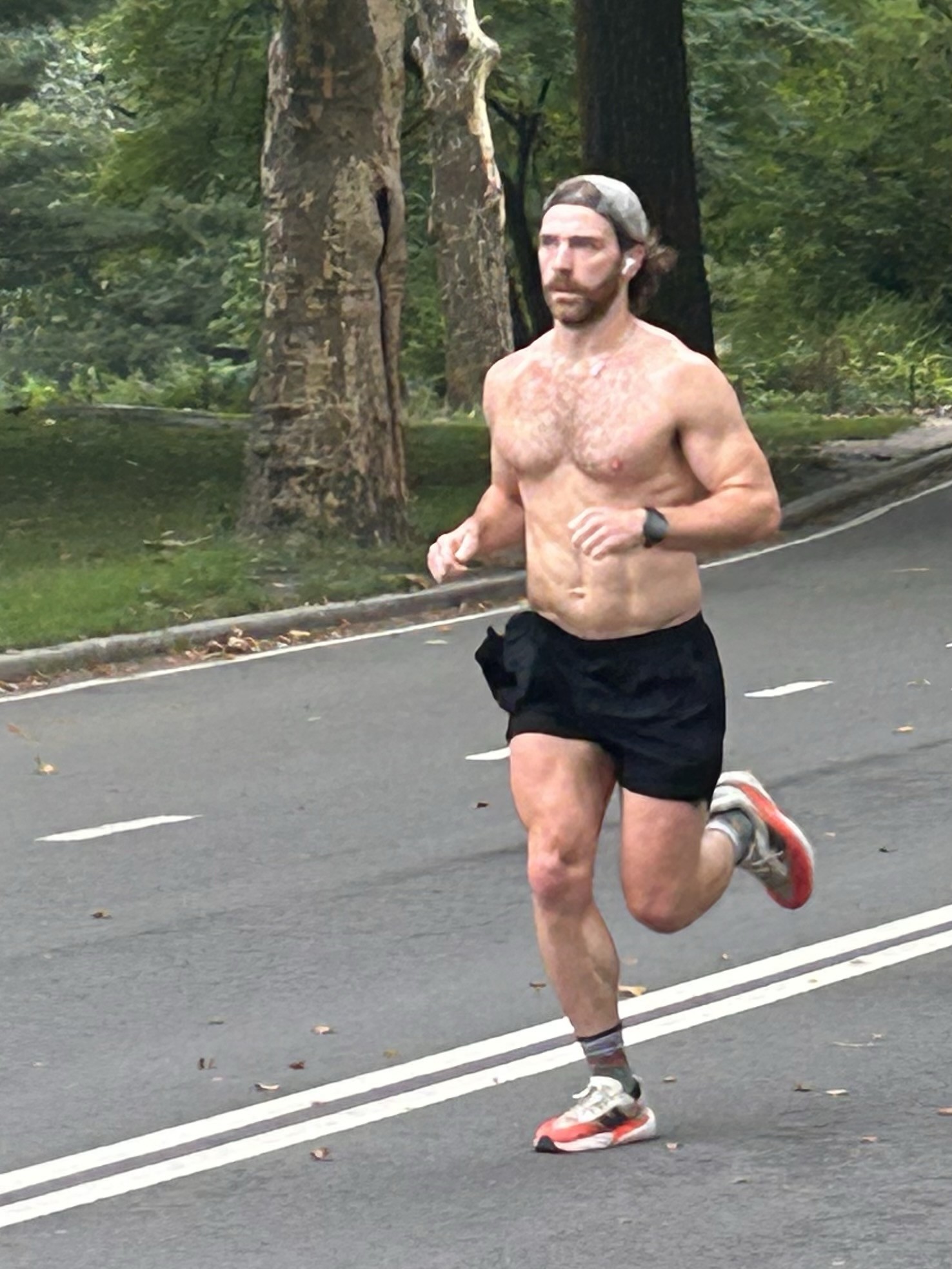 A shirtless male runner with a beard is jogging on a tree-lined road. He is wearing black shorts and athletic shoes.